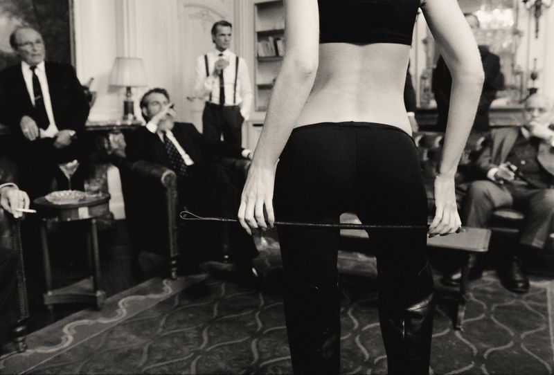Woman in dark crop top and fitted pants holding a riding crop in a room with seated men in suits