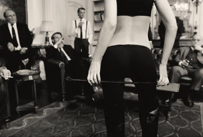 Woman in dark crop top and fitted pants holding a riding crop in a room with seated men in suits