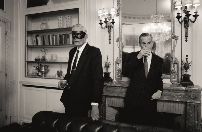 Two men in suits, one wearing a mask, stand indoors with drinks in a black and white photograph. A fireplace and bookshelf are visible.