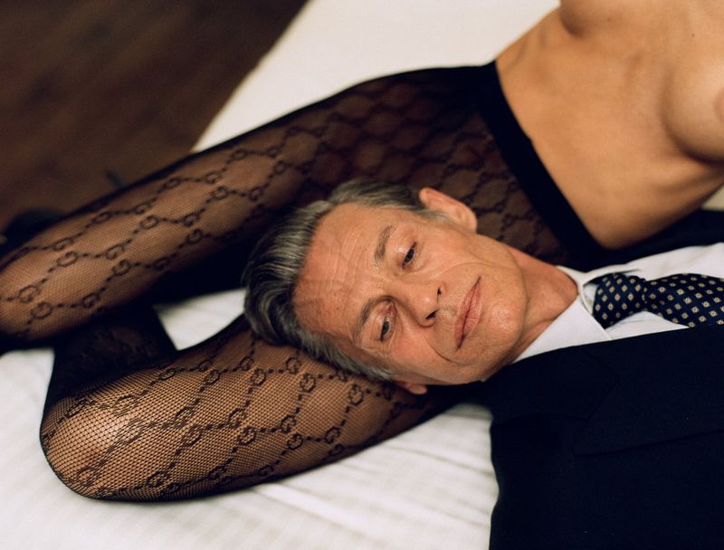 Man in a dark suit with head resting on legs wearing patterned hosiery in a bedroom setting.