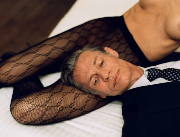 Man in a dark suit with head resting on legs wearing patterned hosiery in a bedroom setting.