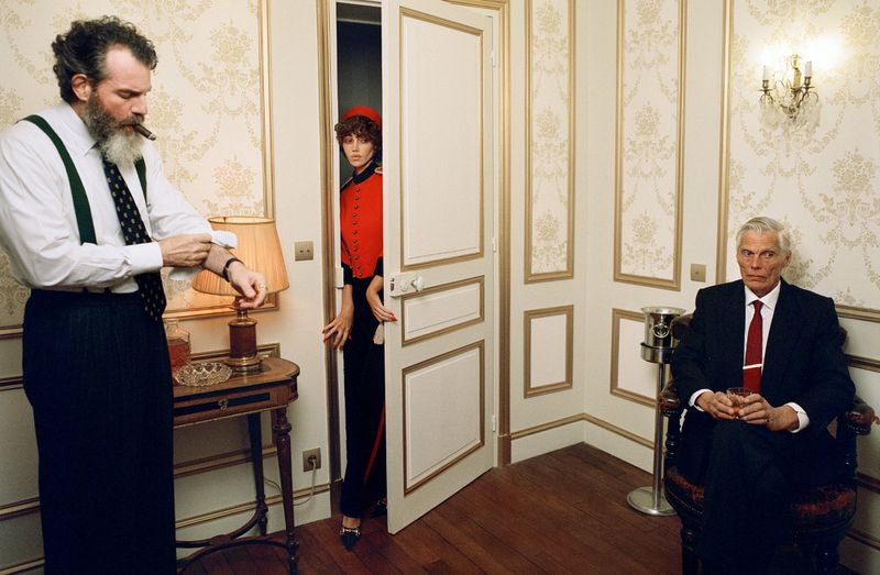 Man with beard adjusting cuff near lamp, woman peeking through a doorway, and seated older man holding a glass in a classic hotel room interior