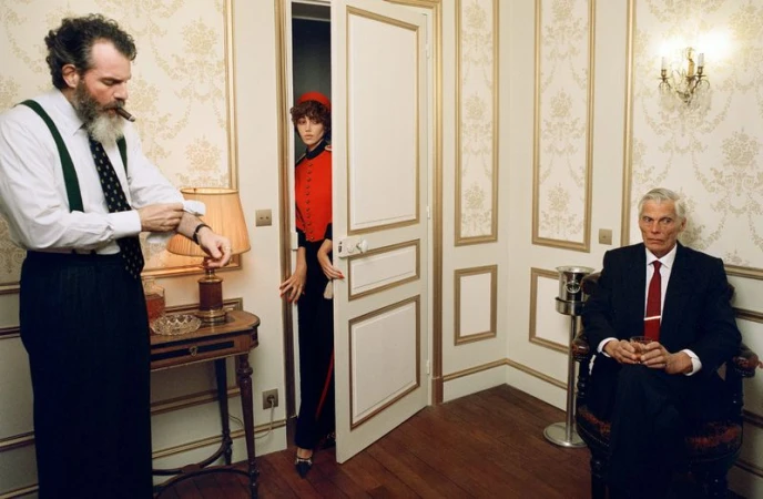 Man with beard adjusting cuff near lamp, woman peeking through a doorway, and seated older man holding a glass in a classic hotel room interior