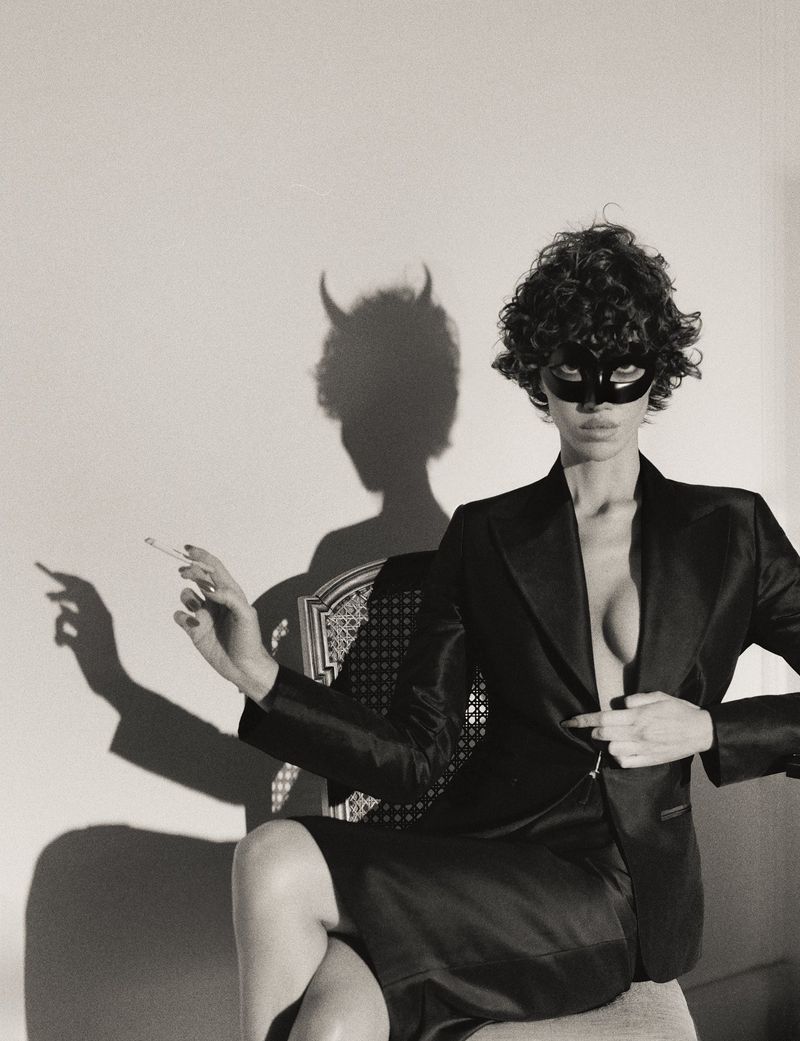 Model wearing a black domino mask and blazer seated on a chair with a dramatic devil-horn shadow on the wall