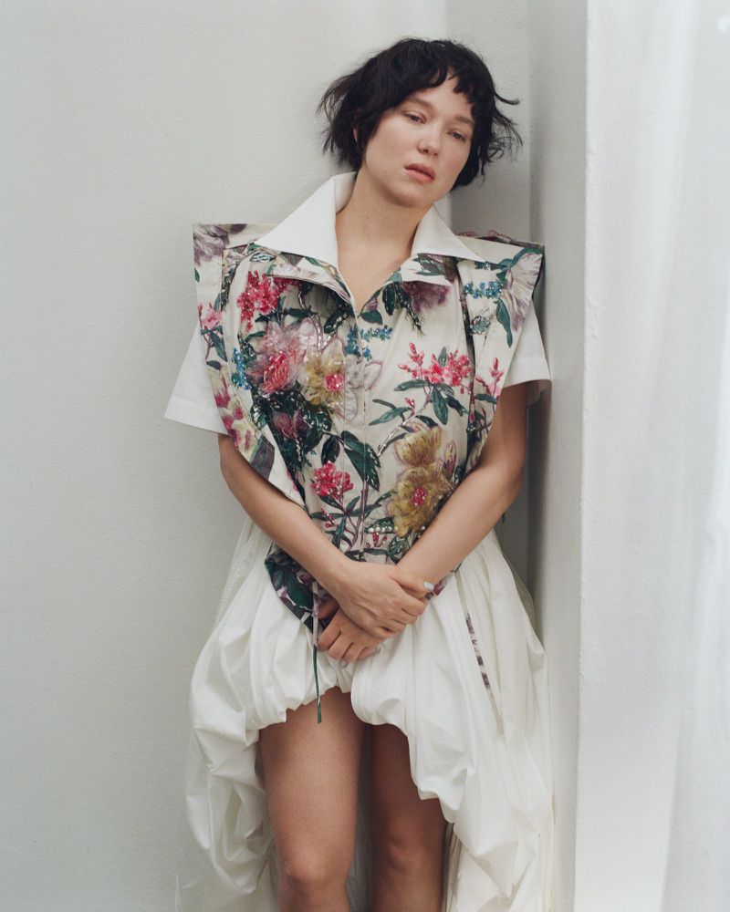 French actress Lea Seydoux posing in a structured floral patterned top and white dress against a plain neutral corner wall.