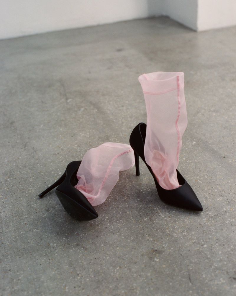 Black stiletto high heels with sheer pink mesh socks placed on a grey concrete floor in a minimalist setting.