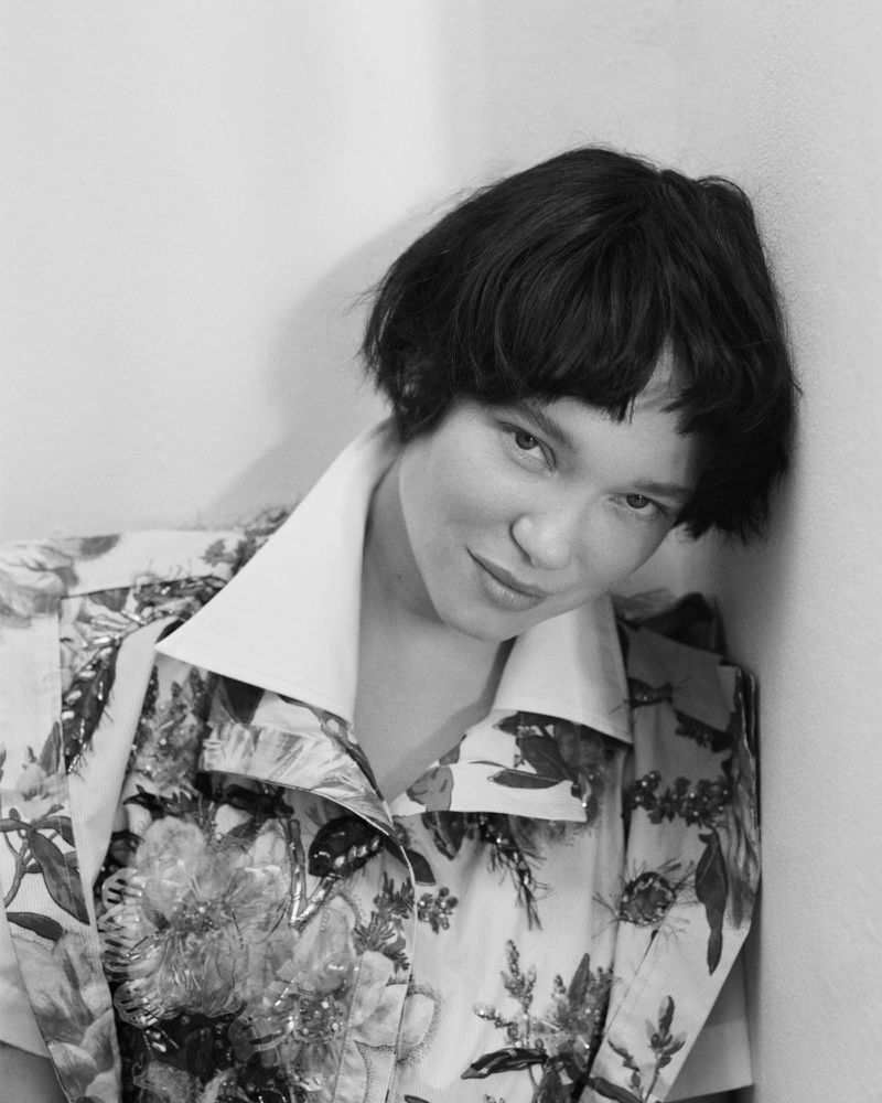 Portrait of actor Lea Seydoux wearing a patterned blouse, leaning against a pale plain wall