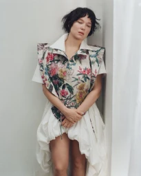 French actress Lea Seydoux posing in a structured floral patterned top and white dress against a plain neutral corner wall.