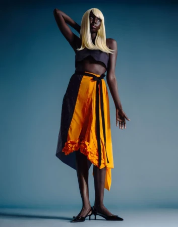 Model wearing a blonde wig, crop top, and yellow wrap skirt standing against a solid blue background in a fashion editorial shot.