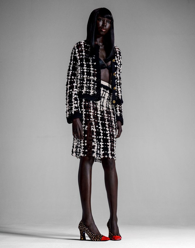 Model Ajok Madel wearing a black and white tweed jacket and matching skirt against a grey seamless studio background.