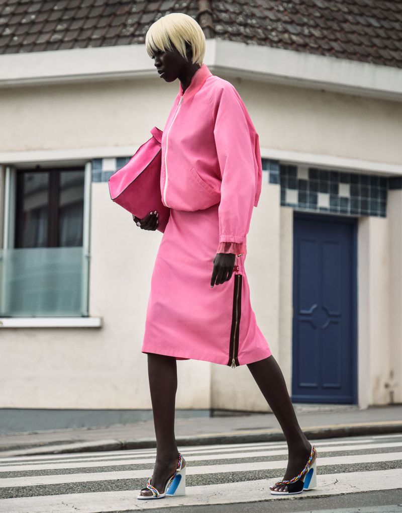 Model wearing a pink jacket and matching skirt holding a pink bag while walking past a building exterior.