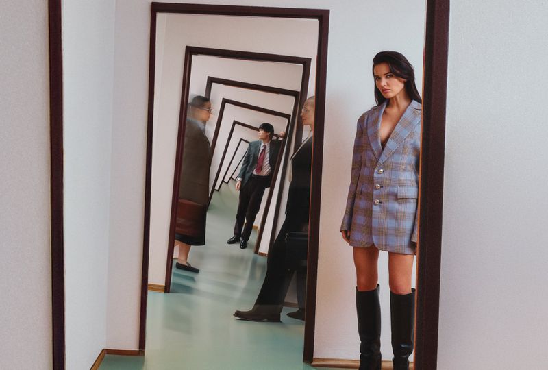 Model wearing a plaid blazer and black knee-high boots standing inside a hallway with multiple receding mirror frames
