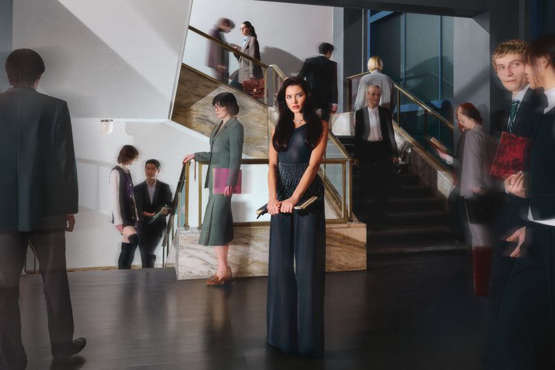 Woman in a dark blue sleeveless jumpsuit standing in a hotel lobby with blurred figures walking on a staircase behind her