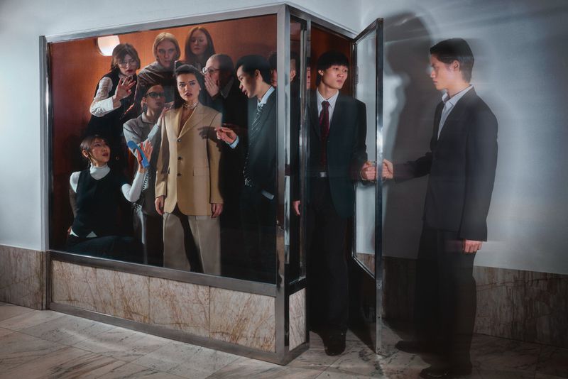 Group of diverse people posed tightly inside a glass display case with one man standing outside holding the door handle
