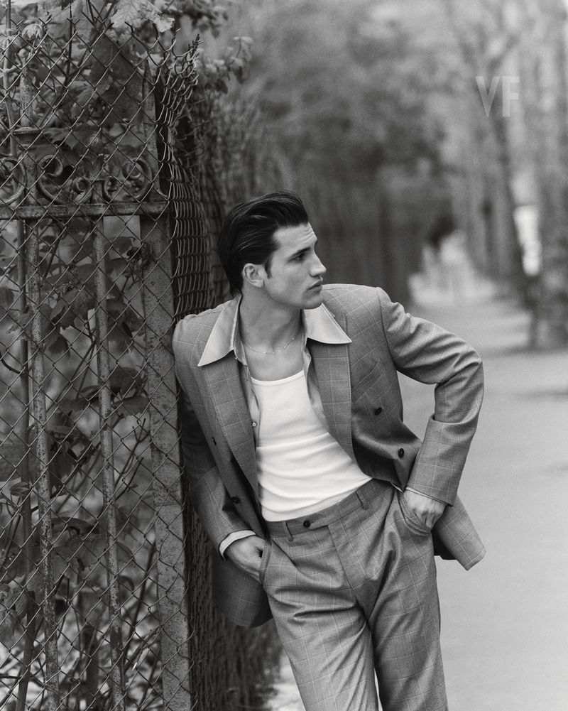 Man wearing a tailored suit jacket over a white tank top leaning against a wire fence on a street