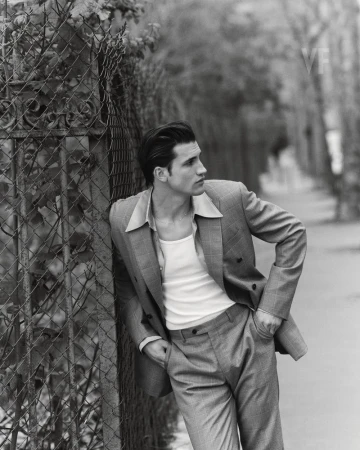 Man wearing a tailored suit jacket over a white tank top leaning against a wire fence on a street