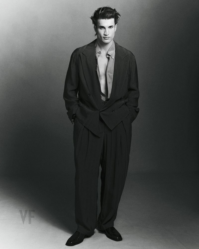 Monochrome full-length portrait of a male model wearing an oversized dark suit jacket and trousers against a gradient studio background.