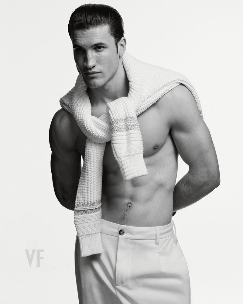 Shirtless male model wearing white trousers and a knitted sweater draped over his shoulders against a plain background.