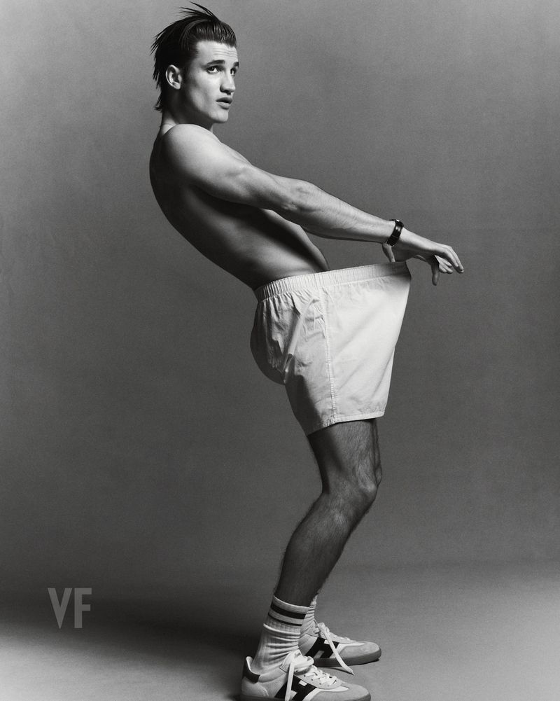Monochrome portrait of a male model wearing white shorts and sneakers, posed in profile against a grey seamless background