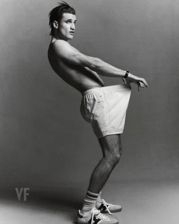 Monochrome portrait of a male model wearing white shorts and sneakers, posed in profile against a grey seamless background
