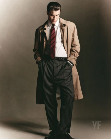 Model wearing a tan trench coat, white shirt, patterned tie, and pinstripe trousers against a neutral background