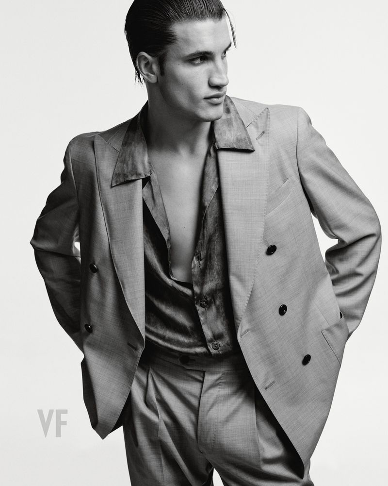 Man wearing a double-breasted grey suit jacket and open shirt against a solid white studio background.