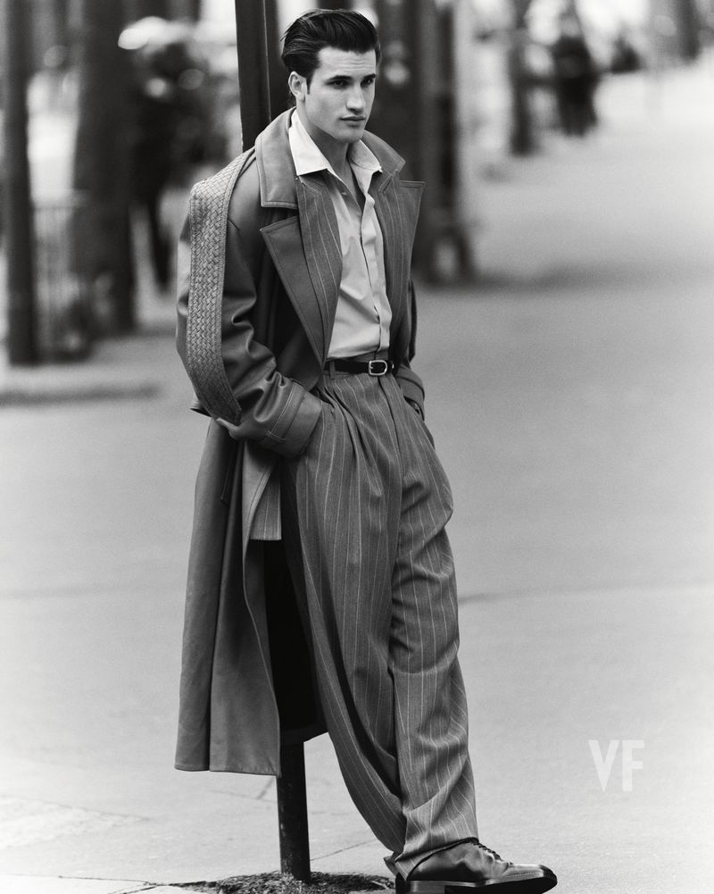 Man wearing a tailored trench coat and trousers leaning against a post on an urban street.