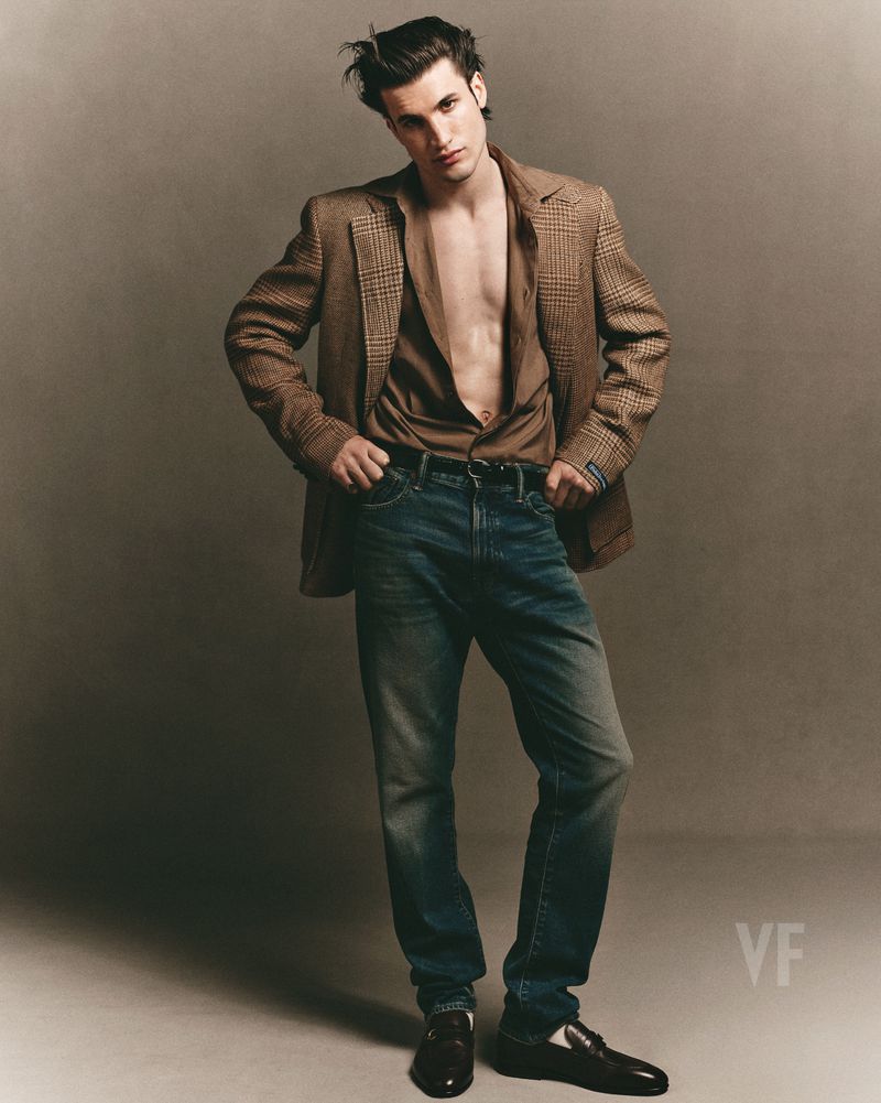 Model Max Knott in a brown plaid blazer and denim jeans posing against a plain grey studio backdrop for Vanity Fair Italia.