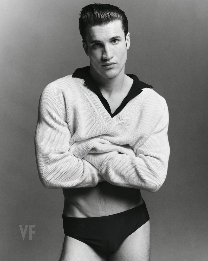 Max Knott posing with arms crossed in a white V-neck sweater and dark briefs against a grey studio seamless backdrop for Vanity Fair Italia.