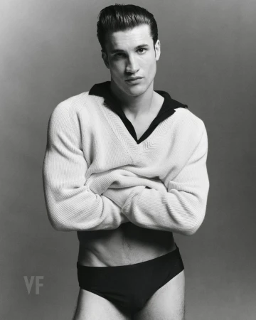 Max Knott posing with arms crossed in a white V-neck sweater and dark briefs against a grey studio seamless backdrop for Vanity Fair Italia.
