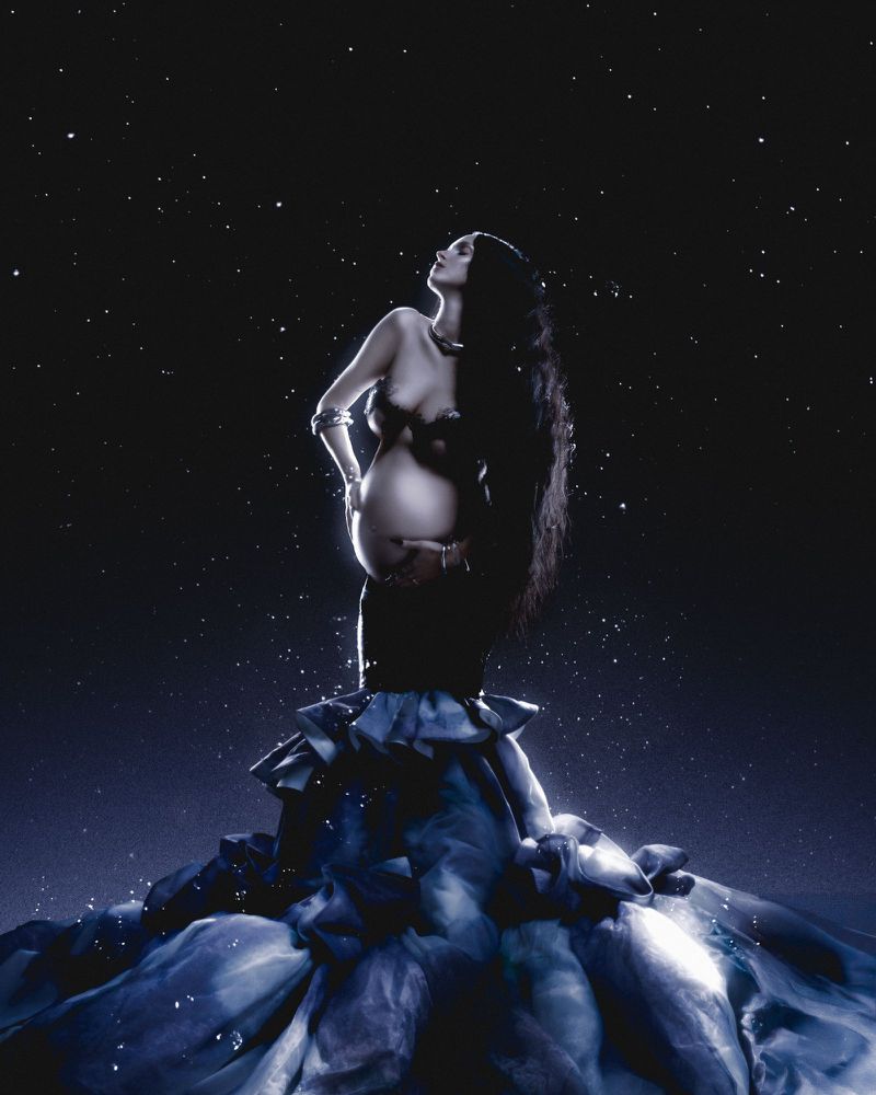 Pregnant woman with long dark hair posing against a starry night sky wearing a dark gown with layered ruffles