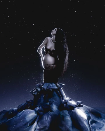 Pregnant woman with long dark hair posing against a starry night sky wearing a dark gown with layered ruffles