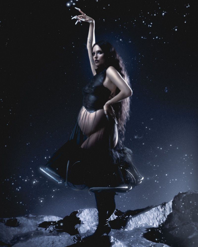 Model with long dark hair wearing a black dress standing on a rocky terrain against a starry night sky