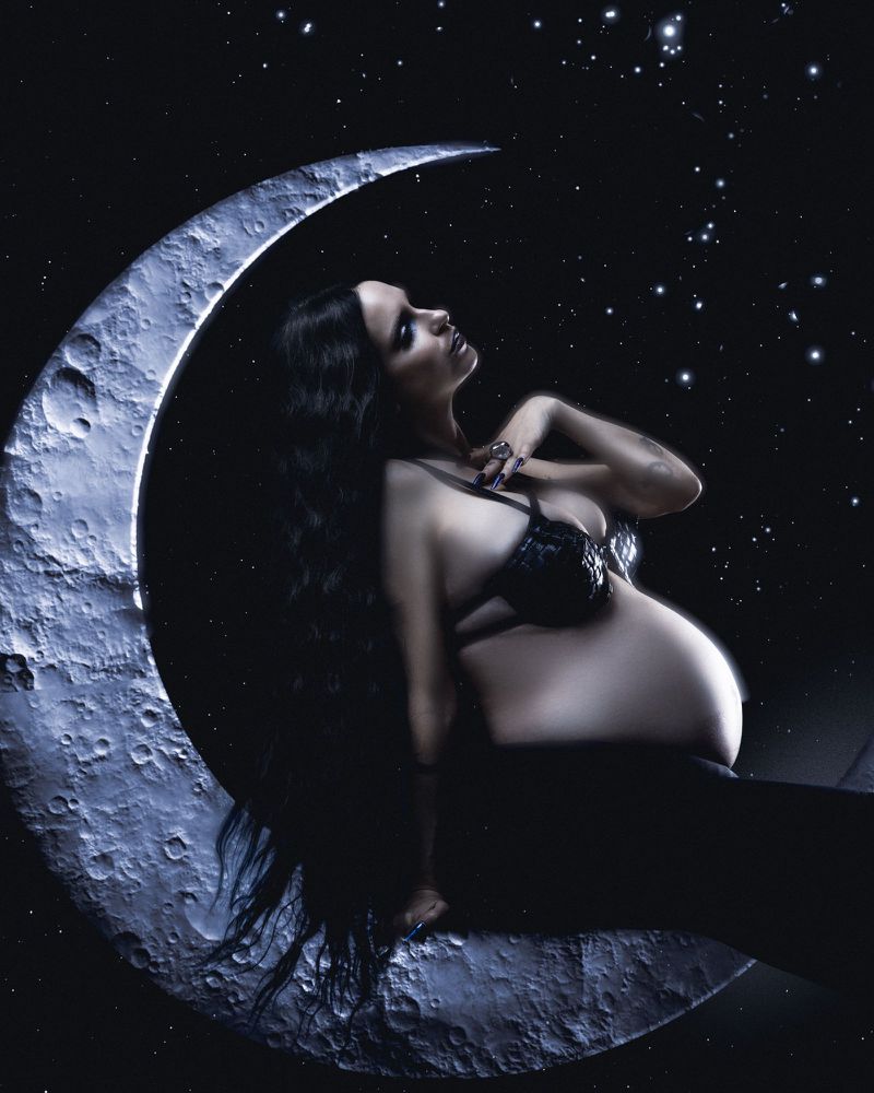 Pregnant woman with long dark hair posing seated on a crescent moon against a star-filled night sky for Kaltblut Magazine editorial