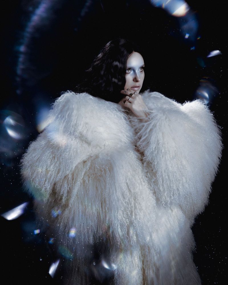 Model wearing a voluminous white faux fur coat against a dark background with floating iridescent light bokeh effects.