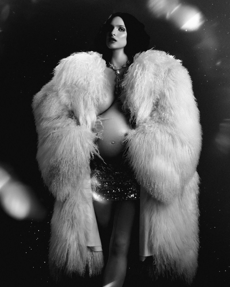 Pregnant woman wearing a large fluffy white fur coat and sequined skirt in a dramatic monochrome fashion editorial portrait.