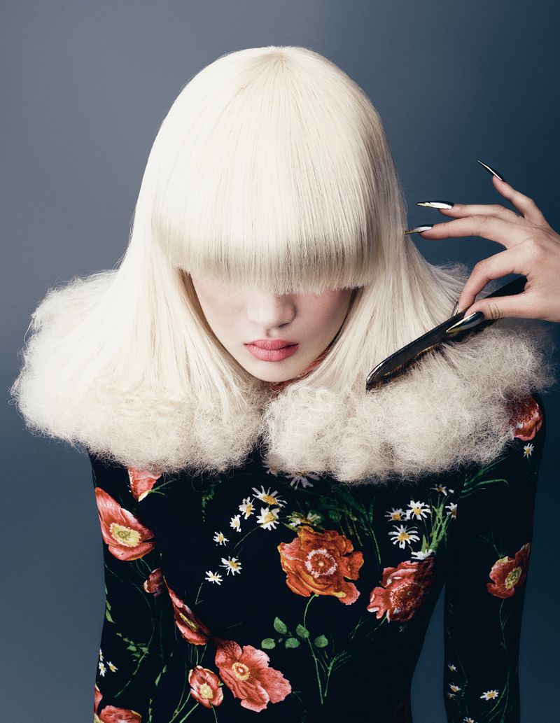 Model with blunt platinum blonde bangs wearing a black floral top, being brushed with long metallic claws.