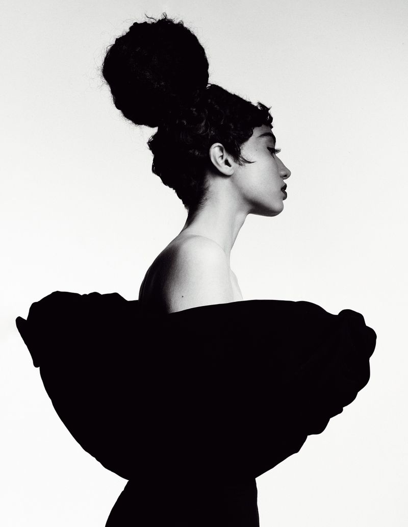 Profile portrait of a model with a voluminous updo hairstyle wearing a sculptural black garment against a white background