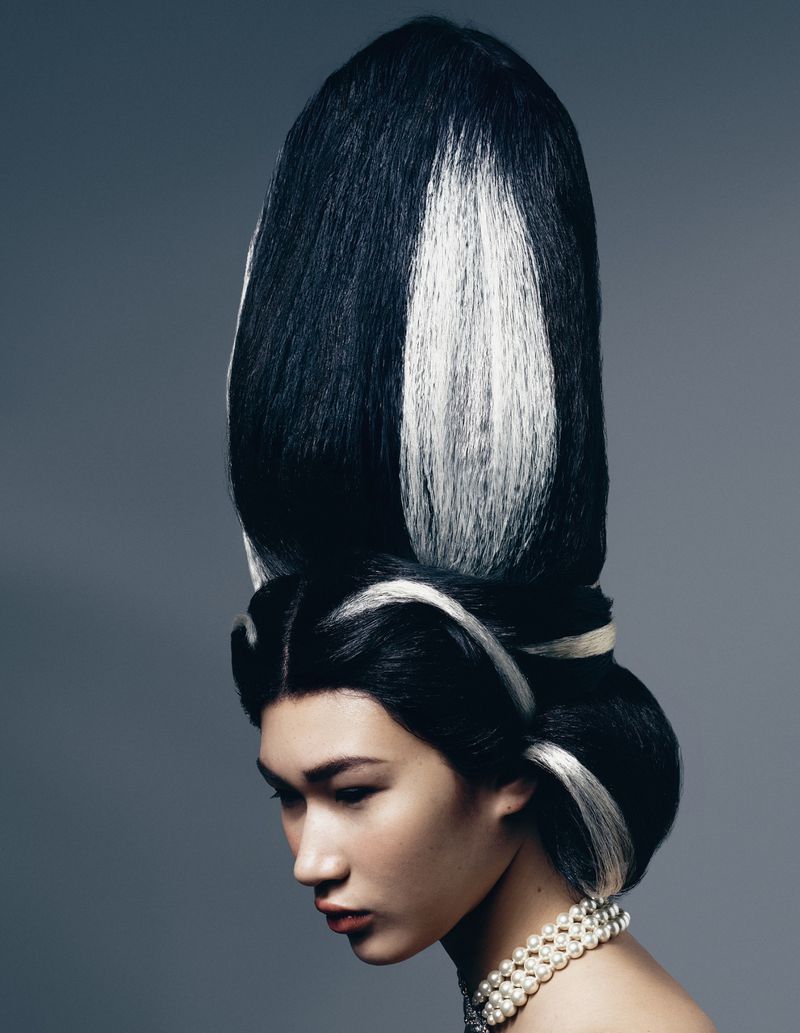 Model with towering black and white sculptural updo hairstyle wearing pearl necklace against plain grey background