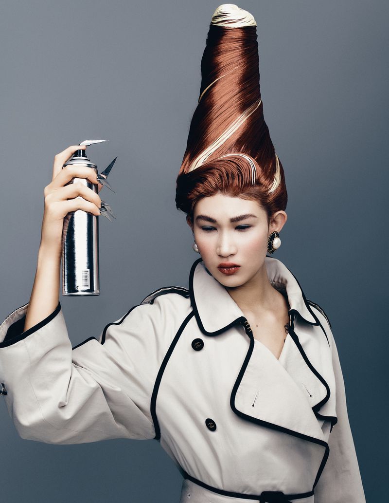 Model with towering sculpted reddish-brown hair holding a silver hairspray can wearing an off-white trench coat against grey background
