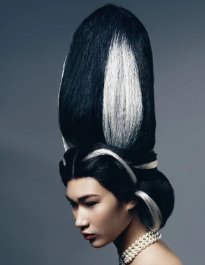 Model with towering black and white sculptural updo hairstyle wearing pearl necklace against plain grey background