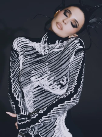 Kylie Jenner wearing a black and white patterned high-neck top with dramatic dark eye makeup against a dark studio background
