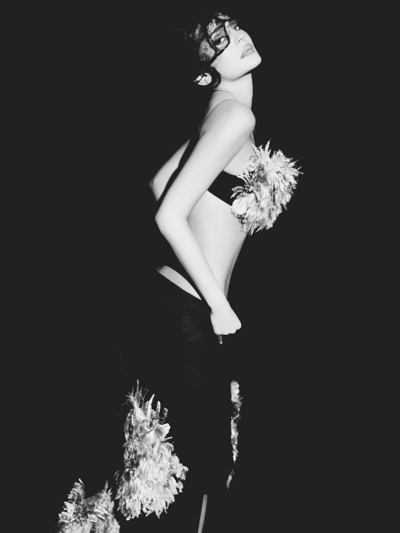 Kylie Jenner in a black lace mask and feathered top against a dark background for Puss Puss Magazine editorial.