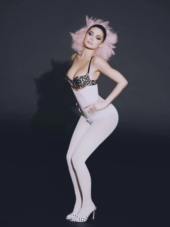 Kylie Jenner with pink hair wearing a lace-trimmed bra and white shapewear bodysuit against a dark background