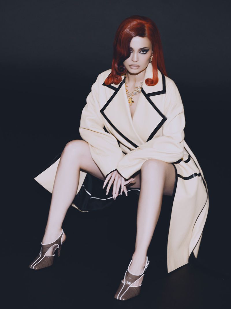 Kylie Jenner with red hair wearing a cream coat with black trim seated against a dark background for Puss Puss Magazine