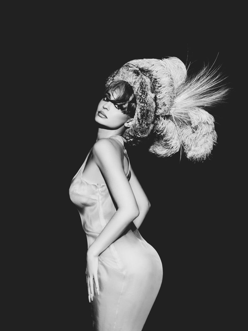 Kylie Jenner wearing a large feathered hat and dress against a dark background, photographed by Kat Irlin for Puss Puss Magazine.