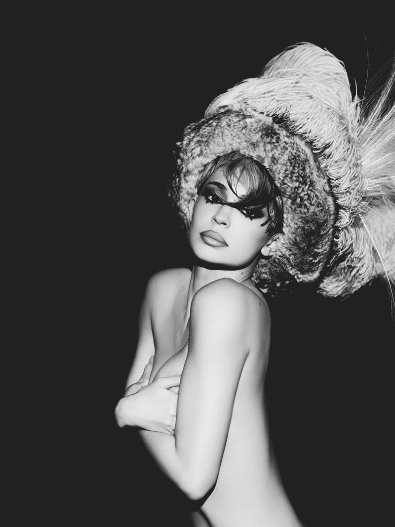 Monochrome portrait of Kylie Jenner wearing an elaborate feathery hat against a dark background for Puss Puss Magazine