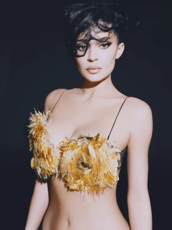 Kylie Jenner wearing a textured yellow feather top with dark wet-look hair against a dark background
