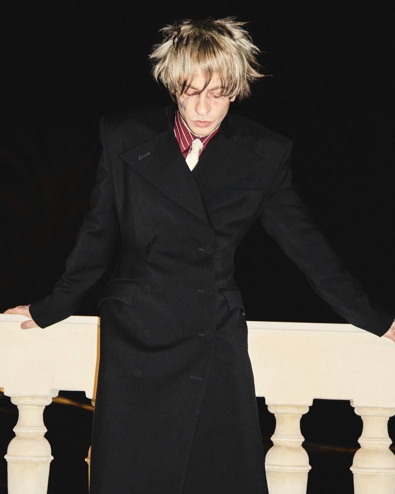 Young man with blonde tousled hair wearing a long black coat against a dark background