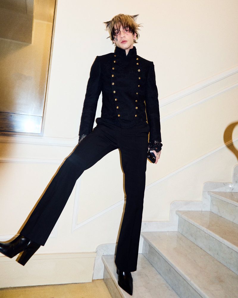 A person in a black suit and boots poses on white marble stairs, with a decorative wall mirror in the background, in an indoor setting.